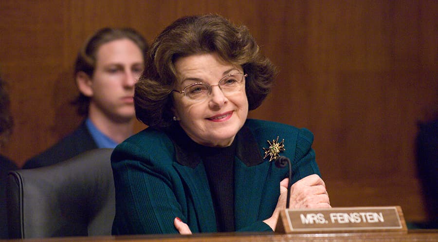 Senator Dianne Feinstein smiling from behind the dais with her "Mrs. Feinstein" name plate in front of her at a Senate Judiciary Committee hearing.