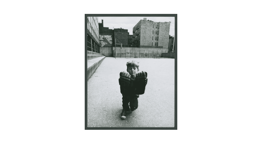 Photograph of a young boy in the middle of an empty lot, kneeling down on one knee with his eyes closed, wearing two hockey gloves.