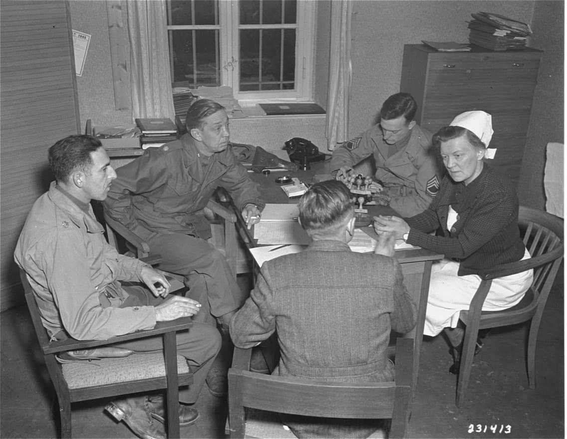 Black and white photo of five people sitting at a table with papers strewn, four leaning over listening, in military uniform, and one is in a nurse's uniform with a hat.